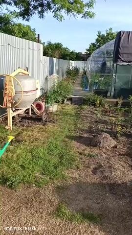 The video opens with a view of a backyard, likely a garden. On the left, there is a cement mixer with yellow and red accents, resting on a patch of dry grass. A corrugated metal fence runs along the left side of the scene, with some greenery behind it. A path, made of what appears to be wooden planks or stepping stones, leads towards a greenhouse in the background. The greenhouse is covered in transparent plastic sheeting and has a dark structure on top. Several plants and bushes are visible along the path and around the greenhouse. The ground is a mix of grass and dirt, with some scattered rocks and debris. The sky is blue with some scattered clouds, suggesting it is daytime. The camera appears to be moving slowly forward, capturing the scene. There are no people clearly visible in the initial frames.