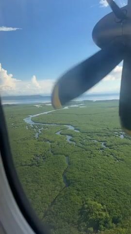 The video presents an aerial view from a small plane, focusing on the landscape below. A large propeller, partially in view through the plane's window, spins against a backdrop of lush, green foliage intersected by winding waterways. Further in the distance, the blue ocean meets the horizon under a bright sky dotted with fluffy white clouds. The overall impression is one of a tropical, river delta region.