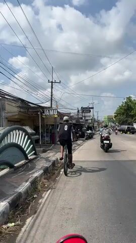 A young person wearing a black vest with the number 0 on the back is riding a bicycle on a paved road. The person is facing away from the camera and appears to be in motion, with the bicycle’s wheels spinning. The setting appears to be a street in a populated area, with buildings lining the right side of the road. Several power lines crisscross overhead. There are other vehicles, including motorcycles, on the road in the distance. The weather is sunny with some clouds in the sky.