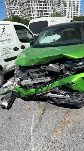A bright green car with significant front-end damage is parked on an asphalt surface. The hood is severely crumpled, and the headlights and front grille are broken and displaced. The windshield is cracked. A white van is parked to the left of the green car. In the background, a tall white building is visible. The scene appears to be a street or parking lot.