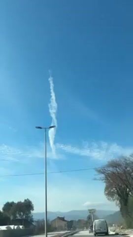 The video shows a view from a car driving on a road. In the clear blue sky, there is a vertical, white, swirling cloud formation. A tall street light is on the left side of the frame, and a few trees are on the right. There are a few cars on the road ahead and buildings in the distance.