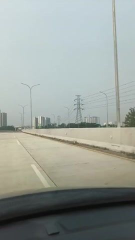 This is a first-person perspective video of a person driving on a highway. The sky is overcast and hazy. On the right side of the highway is a concrete barrier, followed by a field of grass, and then a power line tower with multiple wires stretching across the frame. To the left of the highway are several tall buildings, some of which appear to be under construction or are modern high-rises. Streetlights are also visible along the side of the road. The road itself is paved with concrete and has white lane markings. There are other vehicles in the distance on the highway. The video captures the view from inside a car, showing the dashboard and windshield frame at the bottom of the screen. The overall environment suggests an urban or suburban setting with significant infrastructure.