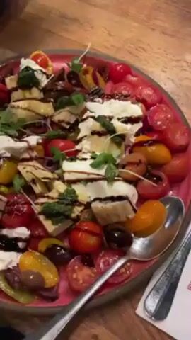 The video shows a dining table with several dishes, including a salad of sliced tomatoes and mozzarella, a flatbread pizza with arugula and balsamic glaze, and a small bowl of salad. The table is made of wood and there is also a glass of liquid with a straw. The camera pans up to show the interior of a restaurant with wooden walls and shelves decorated with plants and bottles. There is also a display case in the background.