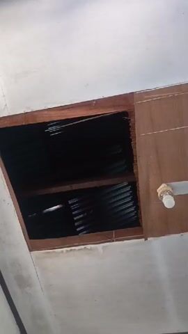 The video shows a close-up view of a ceiling with a large rectangular opening. The opening is framed by wooden planks, some of which appear damaged or unfinished. Inside the opening, a corrugated metal sheet is visible, covering what appears to be the roof structure. The metal sheet is dark and reflective. The ceiling around the opening is painted white. Part of a light fixture is visible on the wooden frame to the right of the opening. The lighting is dim, with some light rays entering through the opening.