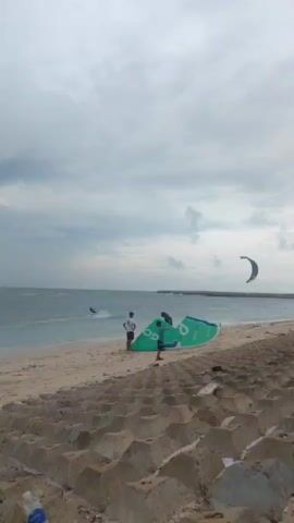 The video captures a daytime scene on a sandy beach, likely in Phú Quý, Vietnam, under an overcast sky. In the foreground, a textured concrete block barrier lines the edge of the beach, with a discarded plastic water bottle visible on it.

On the sandy shore, three individuals are present. Two are standing near a large, green kitesurfing kite laid flat on the sand. This kite prominently features the text "GO" and "VELOCITY GO". A third person is bending over this kite, seemingly adjusting or preparing it.

Further out in the ocean, a kitesurfer is actively riding, leaving a small wake. Two other kites are visible high in the sky; one is a distinct blue and yellow, while another is a darker blue or grey. The ocean appears choppy with small waves, indicative of suitable conditions for kitesurfing. In the distance, a low-lying landmass or a breakwater extends into the sea on the right side of the frame. The overall atmosphere suggests an active watersports location.