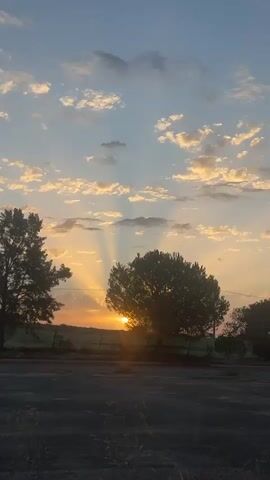 A time-lapse video shows the sun setting over a rural landscape. The sky is a mix of blue and orange, with scattered clouds. The sun is partially obscured by a large tree in the foreground, casting long shadows across the scene. In the distance, there are rolling hills and a fence. The video captures the changing light as the sun sinks below the horizon.