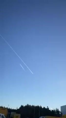 The video shows a bright blue sky with several contrails from airplanes. The contrails appear as straight, white lines stretching across the sky. In the foreground, there is a line of trees and several yellow DHL delivery trucks. The video is taken from a low angle, looking up at the sky.