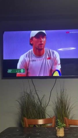 A television displaying a tennis match is mounted on a gray wall. The game is between Ruud and Paul, with the score displayed on the screen. The tennis court is red, and the stands are filled with spectators. Below the television are several faux grass plants in pots, sitting on a dark, speckled table. Two wooden chairs are positioned at the table. Black cords are visible, dangling from the bottom of the television. The lighting is dim.