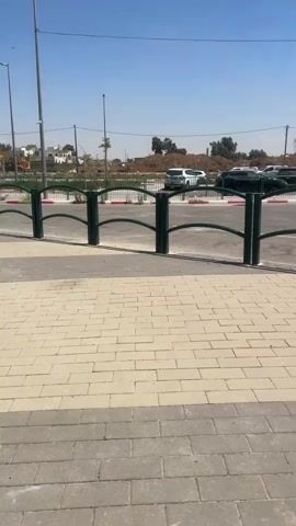 The video captures an outdoor scene on a bright, sunny day. The foreground is dominated by a light-colored brick walkway. A dark green metal fence with curved horizontal bars separates the walkway from a paved area beyond. In the midground, a road or parking lot is visible, with several cars parked or driving. The background features trees, utility poles, and a clear blue sky. The overall impression is that of a public space, possibly a parking lot or street adjacent to a pedestrian area. There are no people visible in the video. The environment appears to be dry and warm, suggesting a summer or dry season. The fence in the foreground is made of two vertical posts and two curved horizontal bars. The paving stones in the foreground are arranged in a pattern. The cars visible in the midground are of various colors and models. The trees are sparse and appear to be of various types, including some that might be palm trees. The sky is clear and blue with a few wispy clouds.