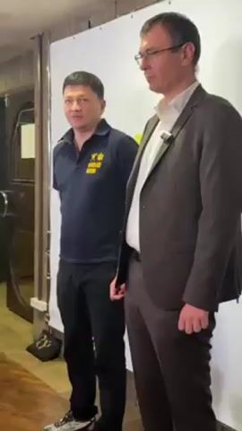 Two men stand in front of a white banner with a yellow design. The man on the left wears a dark blue polo shirt with yellow lettering and a small Ukrainian flag emblem. He has short, dark hair and wears a watch. He is speaking and gesturing with his hands. The man on the right wears a brown suit jacket, a white collared shirt, and glasses. He has short, graying hair. He stands with his arms straight at his sides, holding a small black object in his left hand. A microphone is clipped to his shirt. The backdrop behind them appears to be a wooden wall with decorative elements. A partially visible wooden door is behind the man on the left.