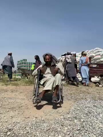 The video captures an outdoor scene under a clear blue sky, likely in Kandahar, Afghanistan, during daylight hours. In the foreground, a man is seated centrally in a manual wheelchair, looking towards the camera with a neutral expression. He wears a light-colored long-sleeved tunic, light trousers, and dark sandals, with a dark turban covering his head. The ground around him is dusty and uneven, covered in gravel.

In the midground and background, several other men are visible, dressed in traditional Afghan attire, including shalwar kameez and various head coverings. They appear to be actively engaged, either standing or moving. To the left, there are large stacks of green boxes or crates, possibly containing aid supplies. To the right, massive piles of white sacks or bags, likely holding commodities such as flour or other food items, are stacked high; some appear to be on the back of a vehicle. The scene suggests a busy distribution point or a gathering where goods are being handled, possibly for aid or commerce, in an open, unpaved area. No discernible text is visible on any objects.