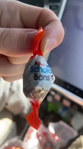 A close-up shot captures a person's thumb and index finger holding a small, individually wrapped chocolate candy. The wrapper is a shiny, crinkled orange plastic twisted at both ends, encasing a foil-like inner wrapper that displays the "Kinder Schoko Bons" branding in blue and white lettering. The chocolate itself is not visible through the opaque part of the wrapper. In the blurred background, what appears to be a white tablet or screen is visible to the right, and other similar wrapped candies are present in the lower portion of the frame. The lighting suggests an indoor or possibly car interior setting.