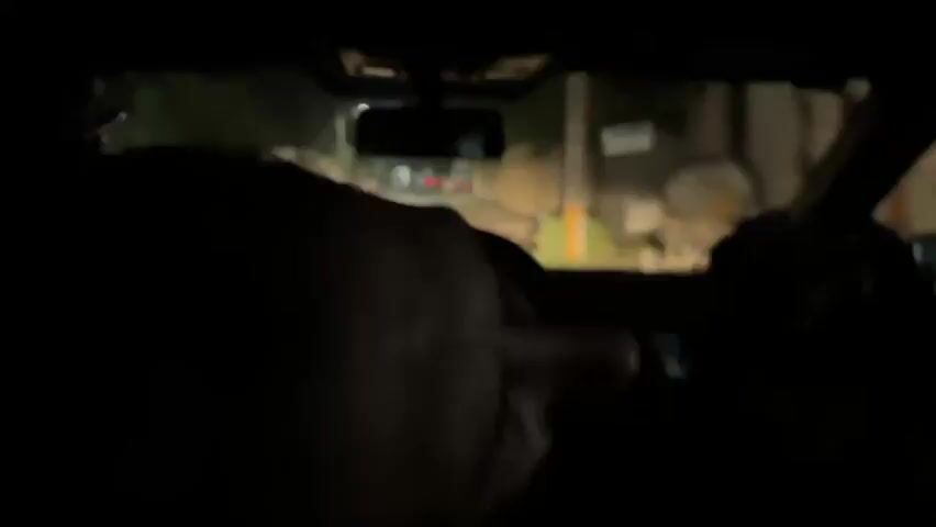 The camera is inside a car driving at night. The interior is dark with the headrests of the front seats partially visible. Outside, the road is lit by headlights. The car passes a wall covered in graffiti, including the word "Circodolo." The perspective is from the back seat, offering a limited view of the road and surroundings.