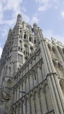 The video shows a low-angle view of a large, ornate stone building, likely a cathedral or historic church. The structure has tall spires, arched windows, and intricate stonework. The camera pans slightly to the right, revealing more of the building's exterior and a small section of the sky with scattered clouds. In the foreground, there are two information boards with maps and text, suggesting this is a tourist site. A low stone wall and a patch of green grass are also visible. The overall impression is one of grandeur and historical significance.