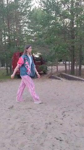 A woman in a denim vest and pink patterned pants walks across a sandy area. She is in a wooded area with pine trees. A red van is parked to her left, and a fire pit constructed of logs is in the foreground.