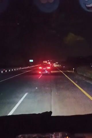 The video shows a nighttime view from the interior of a vehicle, looking out at a highway. Headlights from the vehicle in front illuminate the road ahead, casting bright white beams. A solid yellow line divides the lanes of the dark asphalt highway. In the distance, multiple vehicles are visible, with their red taillights creating streaks of light. Some of these vehicles appear to be stationary or moving slowly, indicated by the concentration of red lights. There are roadside barriers on the left, and sparse vegetation or dark shapes on the right. In the upper part of the frame, decorative, circular objects with cartoon faces are attached to the interior ceiling of the vehicle.