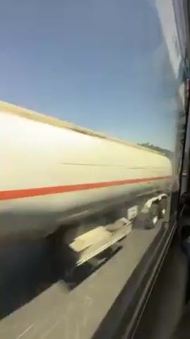 The video, shot from the window of a moving vehicle, shows a road bordered by reddish-brown earth and green trees. A white minivan passes in the opposite direction. The sky is clear and blue. The video is somewhat shaky due to the movement of the vehicle.