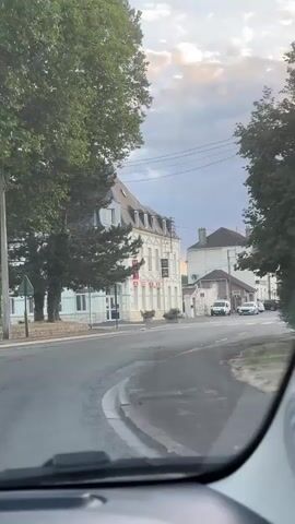 The video appears to be shot from inside a vehicle moving through a town. The time of day is likely late afternoon or early evening, given the lighting and cloudy sky. The road is paved and appears to be a two-lane street with a sidewalk or shoulder on the right side.

On the left side of the road, there is a building with white and grey facades, a mansard roof, and what appears to be commercial signage. Adjacent to this building is a large tree with green foliage.

Further down the road, several cars and vans are parked on the right side. There are also larger vehicles, possibly trucks or delivery vans, parked in a lot or along the roadside. One of these trucks is white with a large green logo.

In the background, the sky is filled with dramatic clouds, with rays of sunlight breaking through in some areas, suggesting a sunset or sunrise. Power lines are visible against the sky. The overall impression is of a quiet, possibly rural or suburban, street scene. There are no people clearly visible in the footage.