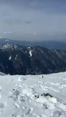 The video shows a panoramic view of a mountainous landscape covered in snow. The foreground is a field of snow with numerous footprints. In the distance, a series of mountain ranges can be seen, some heavily forested and others covered in snow. The sky is overcast with some patches of lighter clouds and glimpses of sunlight. The camera pans across the scene, showcasing the vastness of the snow-covered terrain.