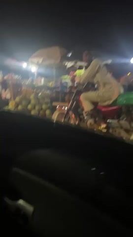 The video, shot at night, depicts a roadside fruit market. The frame is captured from the perspective of a vehicle, likely a car, driving past the scene. Several stalls are visible, each under an umbrella, brightly lit with artificial lights. The stalls display a variety of fruits, including watermelons, pineapples, mangoes, and what appear to be apples. People are present at the stalls, some sitting and others presumably selling or browsing the fruit. A man on a motorcycle is positioned in the foreground, looking toward the camera. The overall atmosphere is that of a bustling nighttime market.