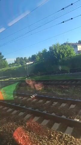A view from inside a moving train shows multiple sets of train tracks stretching into the distance. The tracks are flanked by gravel and green grass. Beyond the tracks, a lush green hillside is dotted with trees and some buildings. The sky is a clear, bright blue with a few wispy clouds and overhead power lines are visible. Sunlight casts a warm glow on the scene.