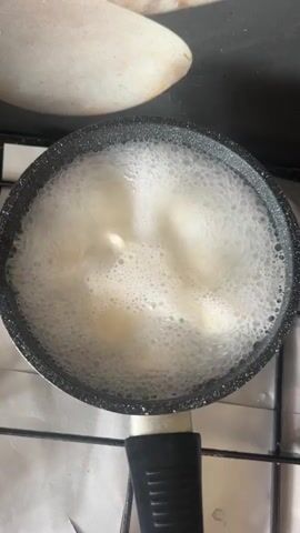 The video shows a top-down view of a small saucepan filled with boiling water. Several pieces of peeled bananas are submerged in the water, bubbling and appearing to soften. The saucepan is black with a metallic rim and a black handle. It sits on a gas stovetop, with the shiny foil of a stovetop liner visible beneath it. A pale, curved object, possibly a countertop or another appliance, is partially visible in the upper portion of the frame. The overall scene depicts the process of boiling bananas.