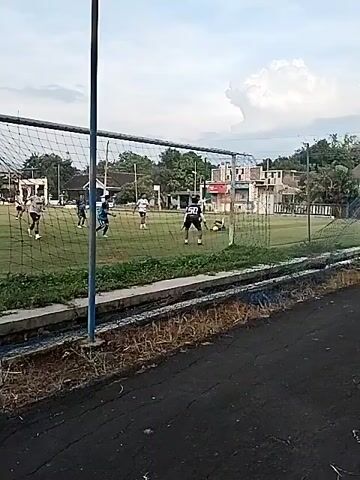 The video shows a soccer game being played on a grassy field.  Several players wearing different colored jerseys can be seen running and moving around the field. A soccer net is visible in the foreground.  A small concrete barrier separates the field from a paved area.  Trees and buildings can be seen in the background under a cloudy sky.  Someone blows a whistle.