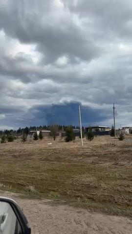 The video captures a wide, rural landscape under a heavily overcast sky. In the foreground, there is a field of dry, light brown grass with scattered green bushes and small coniferous trees. Utility poles supporting power lines are visible in the midground, stretching across the scene. Beyond these, a dense line of trees, including both bare deciduous and evergreen conifers, forms a forested area. Several houses and structures, including a small greenhouse, are nestled among the trees in the distance. The sky is dominated by thick, dark grey and bluish-black clouds, particularly dense and ominous towards the horizon, suggesting an impending storm. The camera slowly pans from left to right, revealing more of this expansive, pre-storm setting.