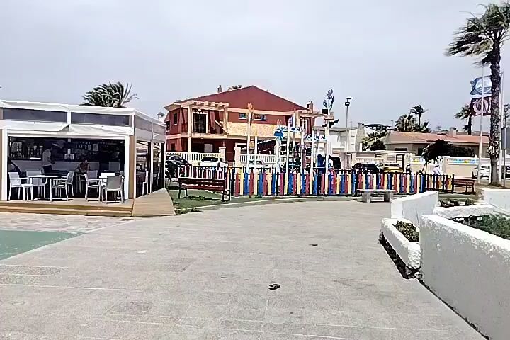 The video shows an outdoor scene at a beach resort on a bright, overcast day. In the foreground, a paved walkway leads to a white, low-lying wall and planters with some vegetation. To the left, there is a white outdoor seating area with tables and chairs, possibly a cafe or restaurant. Beyond this, a playground with colorful fencing and climbing structures is visible. Further back, buildings with red roofs and palm trees line the landscape. The background reveals a sandy beach with several people sunbathing, beach umbrellas, and lounge chairs. The ocean is visible with moderate waves. Wind is noticeable, as indicated by the swaying palm trees. The overall impression is a public area near a beach.