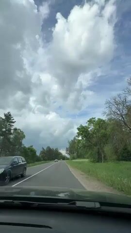 The video depicts a view from the dashboard of a moving vehicle driving on an asphalt road. The sky above is partially covered with billowing white and gray clouds, with patches of blue sky visible. Trees line both sides of the road, primarily green with some deciduous varieties. Cars are seen driving in the same direction as the camera. The camera remains static, capturing the unfolding view.