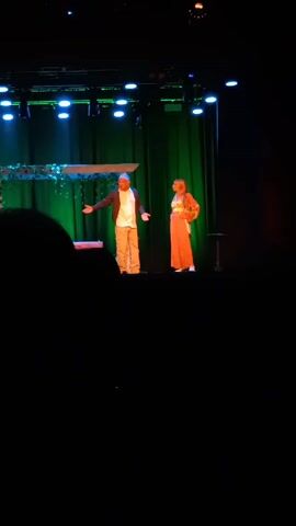 The video appears to capture a live stage performance. Two individuals, seemingly actors, are present on the stage. The male actor is positioned on the left side, clad in a buttoned shirt, jacket, and khaki pants. The female actor is on the right, sporting an orange pair of pants with a patterned crop top. The backdrop is a vibrant green curtain, and above the stage, there's a structure embellished with plants. The stage itself is illuminated by a series of lights. A partial view of an audience member's head is visible in the foreground, suggesting the video was recorded from the audience. The audio captures dialogue, enhancing the contextual understanding of the scene.