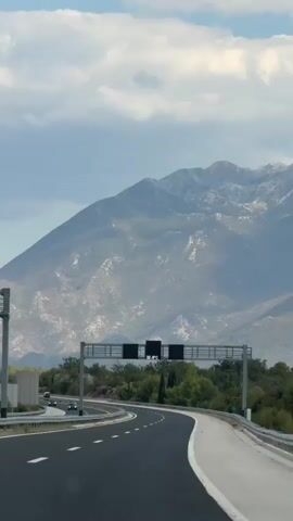 A highway scene is captured from a moving vehicle. The road curves to the right, with clear white lane markings. On the left side of the road, a metal signpost with two dark rectangular panels is visible. The left panel displays a temperature reading of 32.9°C. Beyond the road, lush green trees and shrubs line the landscape. In the background, a large, majestic mountain range rises, its peaks dusted with snow. The sky is partly cloudy, with fluffy white clouds scattered across a pale blue expanse. A few cars are visible on the highway in the distance.