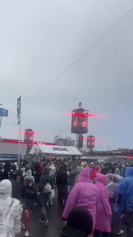 A large crowd of people is gathered outdoors under an overcast sky. Many individuals are wearing rain ponchos in various colors including pink, blue, yellow, green, clear, and black. The ground appears wet. Multiple tall structures with large vertical LED screens are visible, displaying images and illuminated with horizontal red light bars. A main stage area is discernible in the distance. A tall metal pole is present in the foreground at one point, and a food stall or booth labeled "KÄSE-BREZEL" is briefly seen. People are generally walking and standing, facing towards the screens and performance area. Music is playing in the background.