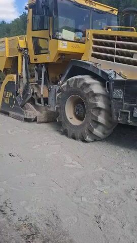 A large yellow machine with large tires is shown operating on a dirt road. The machine has a cutting mechanism at the front which is actively processing the dirt. The machine has "FLEX MIX" written on its side. Another smaller white machine is visible in the background, also on the dirt road. The environment appears to be outdoors with trees and a blue sky with some clouds.
