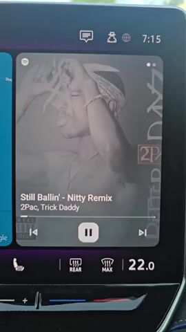 A close-up shot of a car's infotainment system displays album art for the song "Still Ballin' - Nitty Remix" by 2Pac and Trick Daddy. The album art features a black and white image of Tupac Shakur smoking. Below the artwork, playback controls for the song are visible, with the pause button highlighted. The time displayed on the screen is 7:15. Below the music interface, climate control options are visible, including rear defrost, max defrost, and the current temperature set at 22.0 degrees Celsius.