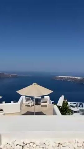 The video shows a scenic view of Santorini, Greece, from a white-walled patio. In the foreground, there's a beige umbrella shading two white chairs. Further out, a white railing surrounds a dining area with more chairs and tables. In the distance, the deep blue Aegean Sea stretches out, dotted with islands. The sky is a clear, bright blue.