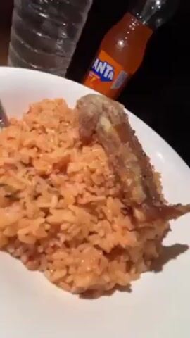 A close-up shot shows a white plate containing a serving of jollof rice, topped with a single fried chicken wing. Behind the plate, out of focus, are a bottle of water labeled "AQUAVIE" and a glass bottle of Fanta orange soda. The background is dark.