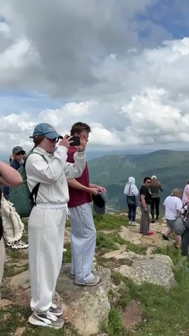 The video shows a group of people standing on a rocky, grassy overlook with a panoramic view of lush green mountains and a cloudy sky. The people are dressed casually, and some are taking photos with their phones. The camera pans across the landscape, highlighting the vastness of the mountains.