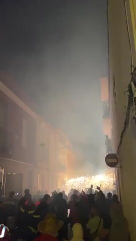 The video shows a nighttime street celebration with a crowd of people gathered in a narrow street lined with buildings. Fireworks are being set off, creating a bright, fiery display in the center of the street, accompanied by a significant amount of smoke that fills the air. Some individuals in the crowd are wearing costumes or festive attire, and a few are holding what appear to be sparklers or other handheld fireworks. People are looking towards the fireworks and some are raising their arms. There are balconies on the buildings, and one person is visible on a balcony observing the event. A red and white circular sign with a horizontal bar is mounted on a pole on the right side of the street. The lighting is dim, with the primary illumination coming from the fireworks and the lights from the buildings. The overall atmosphere is one of excitement and festivity.