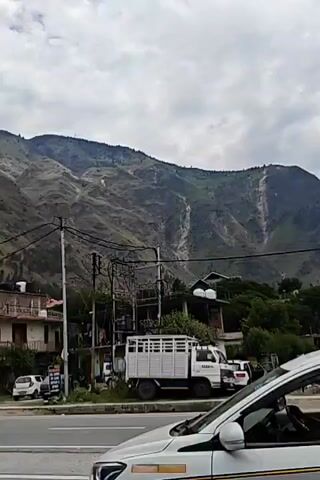 The video showcases a scenic drive through a mountainous region in Jia, India, under a cloudy sky. The camera pans from right to left, revealing a wide road with a central divider. In the foreground, a white Maruti Suzuki Ertiga taxi with a roof rack is seen briefly. Along the roadside, there are multi-story buildings, some appearing to be commercial establishments or homes. Several other vehicles, including cars and vans, are visible on the road and parked along its side. Power lines and utility poles crisscross the frame. The dominant feature of the landscape is a large, rugged mountain range in the background, with patches of green vegetation on its slopes, characteristic of the Himalayan foothills.