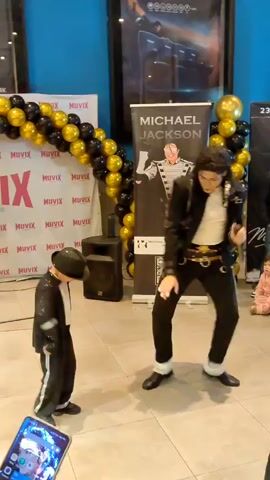 The video captures a lively event, likely a celebration or tribute, in a bright, indoor space. Two individuals, dressed as Michael Jackson, are the focus of the scene. A smaller child, adorned in a miniature MJ costume complete with a hat, energetically mimics the iconic dance moves. Beside him, an adult, also dressed as Michael Jackson, performs with impressive skill and flair.

Behind the performers, a festive atmosphere is evident with a large balloon arch composed of black and gold balloons. Several banners are displayed, one prominently featuring Michael Jackson's name and image, along with a QR code. Another banner displays the Muvix logo. In the background, a movie poster for "MX4D" is visible, suggesting the event might be taking place at a cinema or entertainment venue. A speaker is also present, likely for the music accompanying the performance.

The scene is well-lit, suggesting it's daytime. The overall mood appears joyful and celebratory, with a few spectators, including a young child and adults, observing the performance from the side. The floor is tiled, and the walls are painted in a vibrant blue. The event seems to be well-attended and filled with enthusiasm for the legendary performer.