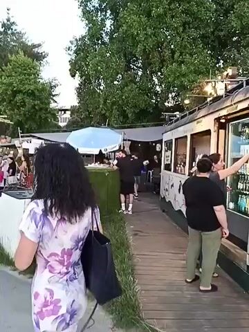 The video shows a bustling outdoor scene, possibly a beachside establishment or festival. In the foreground, a woman with curly dark hair wearing a floral print dress walks away from the camera, carrying a dark tote bag. The path ahead is made of wood planks. To the right, a structure resembling a food or drink stall is visible, with people inside and a counter with what appears to be drinks. This stall has a distinctive green and white design with black paw prints on the lower half. A blue and white umbrella stands near the stall. Further into the scene, there are more stalls, tables, and people milling about. Trees and foliage are prominent throughout the background. In the distance, a modern building can be seen. There are also portable air conditioning units. The lighting suggests it is either late afternoon or early evening. People are seen relaxing on beach chairs in a sandy area. A tall wooden tripod structure with fabric draped over it is present, adding to a bohemian or beach-like atmosphere. A street lamp is also visible. The general impression is that of a lively gathering place.