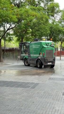 A medium shot captures an urban scene where a green street cleaning vehicle is actively spraying water onto a paved surface. The vehicle, labeled "MATHIEU" and bearing the text "El Prat" and "Netegem els contenidors amb aigua a pressió." (We clean the containers with pressurized water.) in Catalan, moves from right to left, leaving a wet trail behind it. Its license plate "S 0000 QCP" is visible. A man, dressed in a dark green t-shirt, black shorts, and black athletic shoes, walks from right to left in the foreground, holding a phone to his ear. The area is lined with green trees and features a grid-patterned grate in the foreground. The weather appears to be overcast or the scene is under significant shade, and the ground is damp from the cleaning activity.