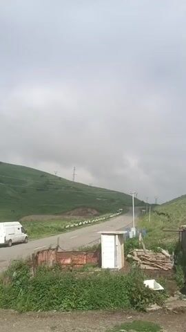 A white van is parked on the side of a paved road. To the left, a green, grassy hill slopes upwards. The sky is overcast with gray clouds. In the foreground, there is some overgrown vegetation and a small, white structure. Further up the hill, there are power lines and poles. The road curves to the right and goes up the hill.
