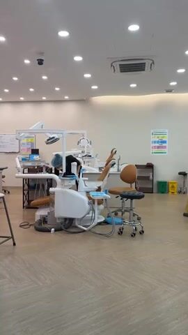 The video shows the interior of a dental clinic. Two individuals are present, both wearing scrubs and surgical masks. The person on the left is wearing blue scrubs and is bent over a dental chair, appearing to be engaged in some task. The person on the right is wearing yellow scrubs and is standing next to a dental chair, adjusting a light fixture. Several dental chairs with attached equipment, as well as sinks and cabinets, are visible. The room is well-lit with ceiling lights. The floor is made of wood-like tiles. There are also bins and posters on the wall. The overall environment suggests a clean and professional medical setting.