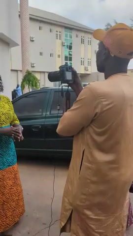 The video captures a scene in Unguwar Sarki, Nigeria. The primary subjects are several individuals, including a woman in a vibrant yellow, turquoise, and orange patterned dress being interviewed by a person holding a video camera. Another woman, dressed in a light-colored hijab and a patterned abaya, is looking at her phone. A man in a light blue traditional outfit is walking in the background.

The setting appears to be an outdoor area in front of a multi-story building, likely a hotel or office complex, given the numerous air conditioning units visible on the facade. Several cars are parked in a lot, including a dark-colored sedan and a red car. The ground is paved, and there is some greenery and shrubbery bordering the parking area. The time of day seems to be daytime, possibly late afternoon, with overcast or soft lighting.

The activity clearly depicts an interview or a media recording session, with the woman in the patterned dress actively engaged in speaking. The overall mood is neutral, focusing on the interaction and the environment. No visible text is present in the video.