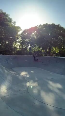A person on a scooter rides in a concrete skate park. The person jumps into the air with the scooter. The person lands and continues to ride. Other people are in the background of the skate park.