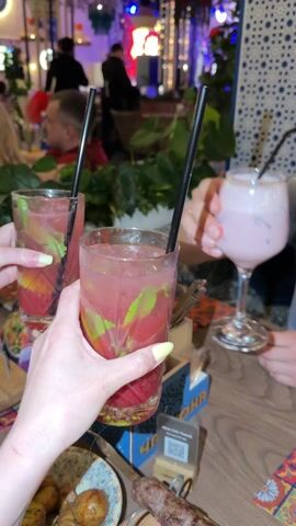 The video shows a toast with three drinks, two red cocktails with mint and one pink cocktail, at a restaurant. The table is covered with colorful tiles. There is a plate of food and a QR code visible. In the background, people are seated and decorations are present.