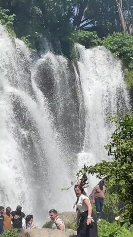 The video shows a large waterfall with several people gathered at its base. The waterfall is surrounded by lush greenery and rocks. The people appear to be tourists, with some posing for photos and others observing the natural beauty. The water is flowing vigorously, creating a misty spray that is visible in the air. The overall impression is one of a scenic and enjoyable outdoor experience.