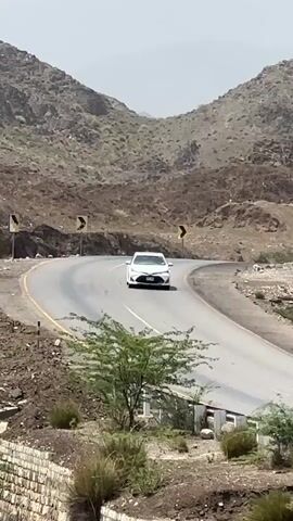 This media file shows a winding road through a mountainous, arid landscape. The road is paved and appears to be in good condition, with yellow directional arrows marking curves. A white sedan is driving on the road, appearing in the foreground of the first few frames before receding into the distance. The surrounding terrain is characterized by rocky, brown hills and sparse vegetation, with some green shrubs and trees. The sky is blue with a few white clouds, suggesting a clear, sunny day. The overall impression is of a remote and rugged location, possibly in a desert or mountainous region. There are no people visible in the media, and the focus is on the natural landscape and the car traversing it. The time of day appears to be daytime, given the lighting. There is no visible text in the media.